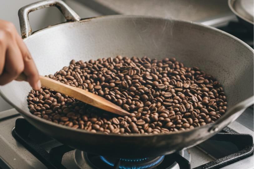 A person using a wooden spatula to roast coffee beans in a large metal wok over a blue gas flame. Light steam or smoke rises from the beans as they are being stirred. A person using a wooden spatula to roast coffee beans in a large metal wok over a blue gas flame. Light steam or smoke rises from the beans as they are being stirred.
