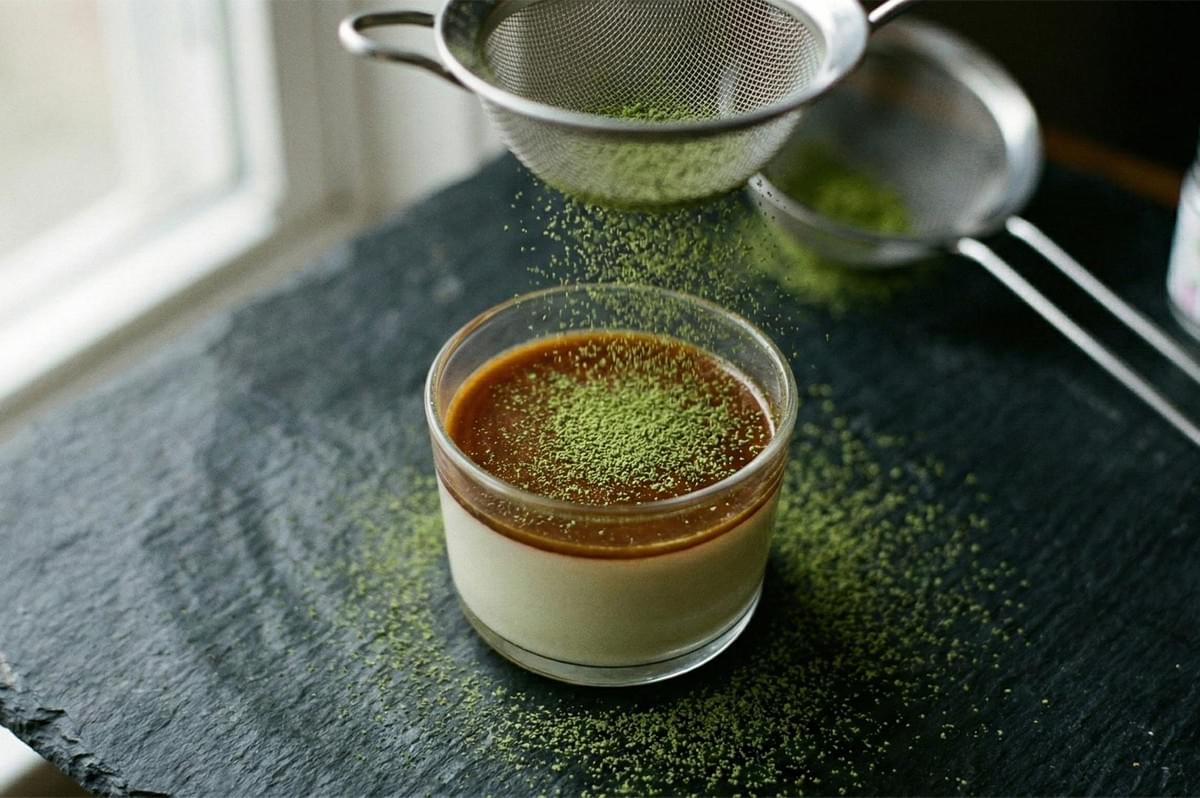 An action shot of bright green matcha powder being sifted through a fine-mesh strainer over a caramel-topped pudding in a glass. An action shot of bright green matcha powder being sifted through a fine-mesh strainer over a caramel-topped pudding in a glass.