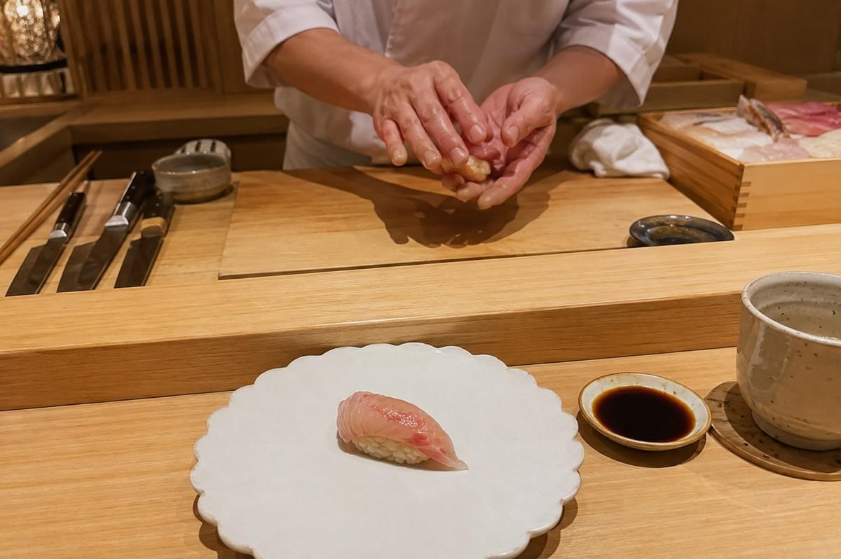 A chef in a white uniform carefully shapes a piece of nigiri sushi with both hands behind a clean, light wooden omakase counter, where a wooden prep board, lined Japanese knives, and a wooden box of raw fish portions sit ready. In the foreground, a scalloped white ceramic plate presents a single piece of fresh sea bream nigiri alongside a small dish of soy sauce and a stone cup, captured from a diner's eye-level perspective in a warm, minimalist Japanese restaurant environment. A chef in a white uniform carefully shapes a piece of nigiri sushi with both hands behind a clean, light wooden omakase counter, where a wooden prep board, lined Japanese knives, and a wooden box of raw fish portions sit ready. In the foreground, a scalloped white ceramic plate presents a single piece of fresh sea bream nigiri alongside a small dish of soy sauce and a stone cup, captured from a diner's eye-level perspective in a warm, minimalist Japanese restaurant environment.