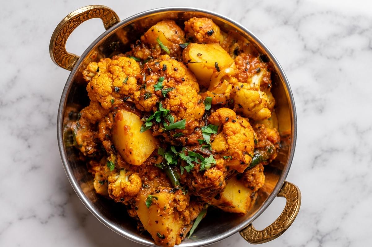 A metal kadai bowl filled with cooked Aloo Gobi garnished with herbs on a marble surface. A metal kadai bowl filled with cooked Aloo Gobi garnished with herbs on a marble surface.
