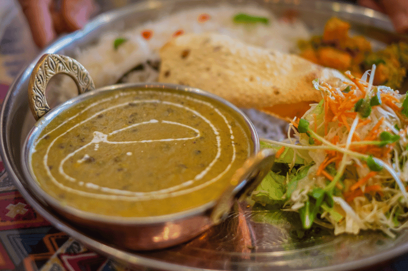 A close-up of a rich, creamy lentil soup (dal) with a white garnish swirl, served in a traditional metal bowl alongside a fresh cabbage and carrot salad and a crispy papadum. A close-up of a rich, creamy lentil soup (dal) with a white garnish swirl, served in a traditional metal bowl alongside a fresh cabbage and carrot salad and a crispy papadum.