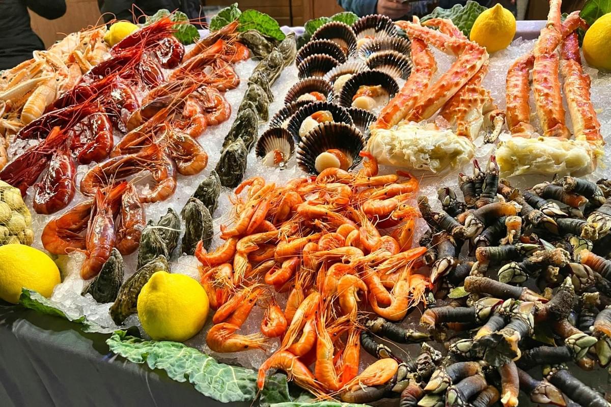 A lavish display of fresh seafood is spread across crushed ice on a table lined with green leaves and whole lemons. The assortment features various types of red and orange shrimp, oysters, scallops in dark shells, thick king crab legs, and dark goose barnacles. A lavish display of fresh seafood is spread across crushed ice on a table lined with green leaves and whole lemons. The assortment features various types of red and orange shrimp, oysters, scallops in dark shells, thick king crab legs, and dark goose barnacles.