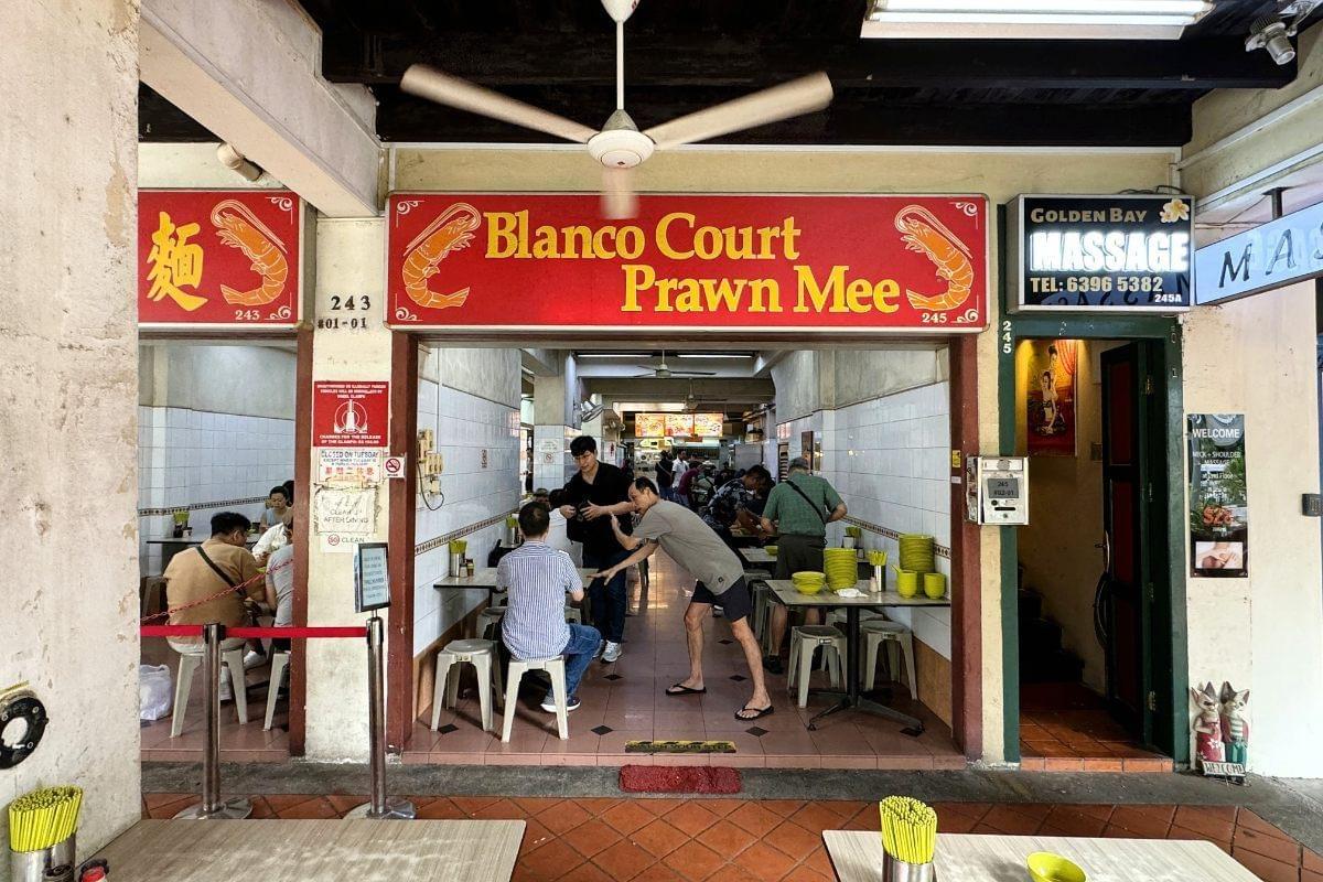 A bright red sign reading "Blanco Court Prawn Mee" hangs over the open entrance of an eatery filled with patrons dining at small tables. Inside, staff members clear dishes and interact with customers, while a ceiling fan spins overhead. A bright red sign reading "Blanco Court Prawn Mee" hangs over the open entrance of an eatery filled with patrons dining at small tables. Inside, staff members clear dishes and interact with customers, while a ceiling fan spins overhead.