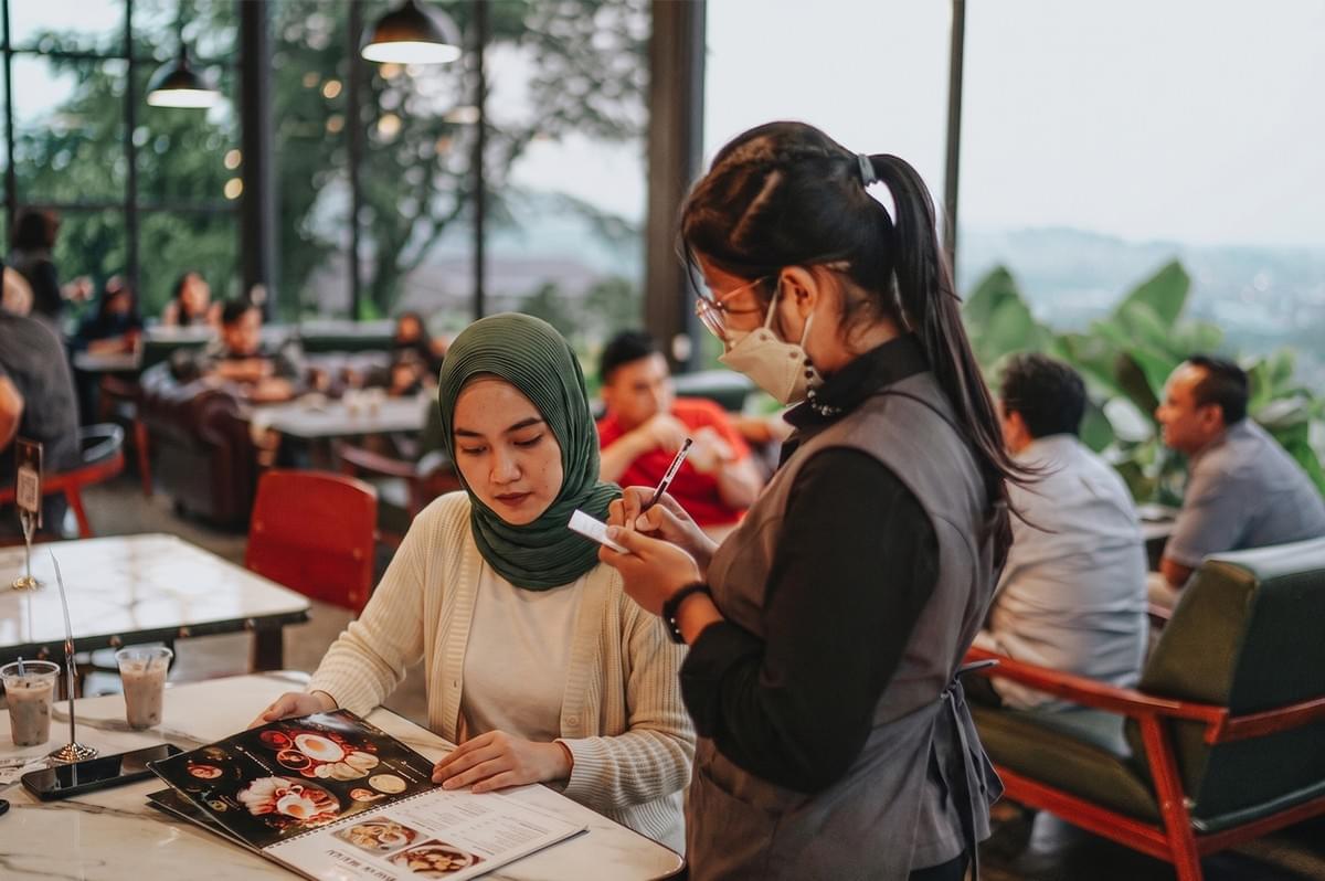 A customer in a green hijab looks at a menu while a server in a face mask takes an order in a busy, modern restaurant with large windows. A customer in a green hijab looks at a menu while a server in a face mask takes an order in a busy, modern restaurant with large windows.