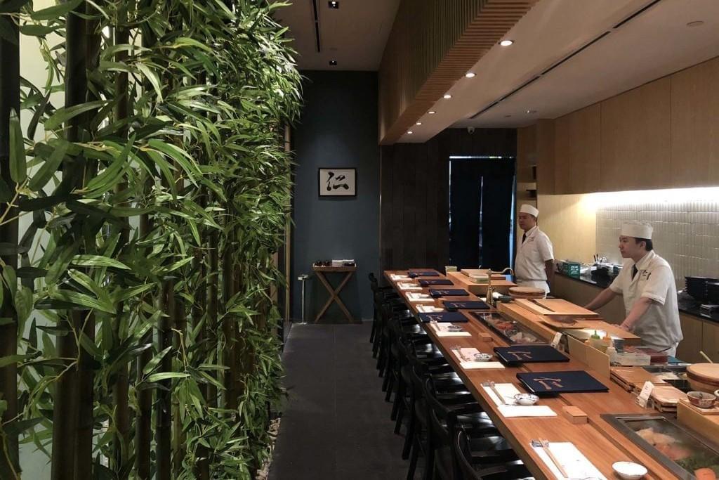 Two sushi chefs stand behind a long, wooden omakase dining counter neatly arranged with place settings and dark chairs. A lush decorative wall of tall green bamboo stalks lines the left side, adding a natural feel to the modern restaurant interior. Two sushi chefs stand behind a long, wooden omakase dining counter neatly arranged with place settings and dark chairs. A lush decorative wall of tall green bamboo stalks lines the left side, adding a natural feel to the modern restaurant interior.