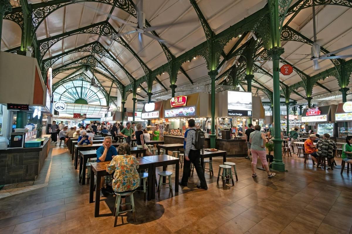 A spacious, indoor food hall features decorative green cast-iron arches and vaulted ceilings above several food stalls. Patrons sit at long wooden dining tables, while others walk between the various hawker stalls displaying bright signage. A spacious, indoor food hall features decorative green cast-iron arches and vaulted ceilings above several food stalls. Patrons sit at long wooden dining tables, while others walk between the various hawker stalls displaying bright signage.