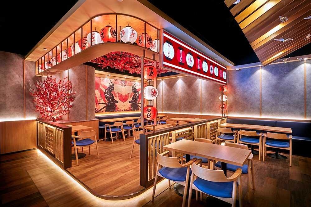 An interior view of a Japanese restaurant features wooden dining tables and chairs with blue cushions, arranged under an architectural canopy decorated with hanging red and white lanterns. The background showcases a vibrant wall mural depicting a samurai, complemented by warm lighting and faux cherry blossom trees. An interior view of a Japanese restaurant features wooden dining tables and chairs with blue cushions, arranged under an architectural canopy decorated with hanging red and white lanterns. The background showcases a vibrant wall mural depicting a samurai, complemented by warm lighting and faux cherry blossom trees.