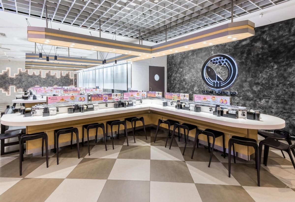 An empty Sushi Express restaurant features a long, modern counter lined with black stools and a conveyor belt system for dishes. A textured grey accent wall displays the illuminated round restaurant logo behind the seating area. An empty Sushi Express restaurant features a long, modern counter lined with black stools and a conveyor belt system for dishes. A textured grey accent wall displays the illuminated round restaurant logo behind the seating area.