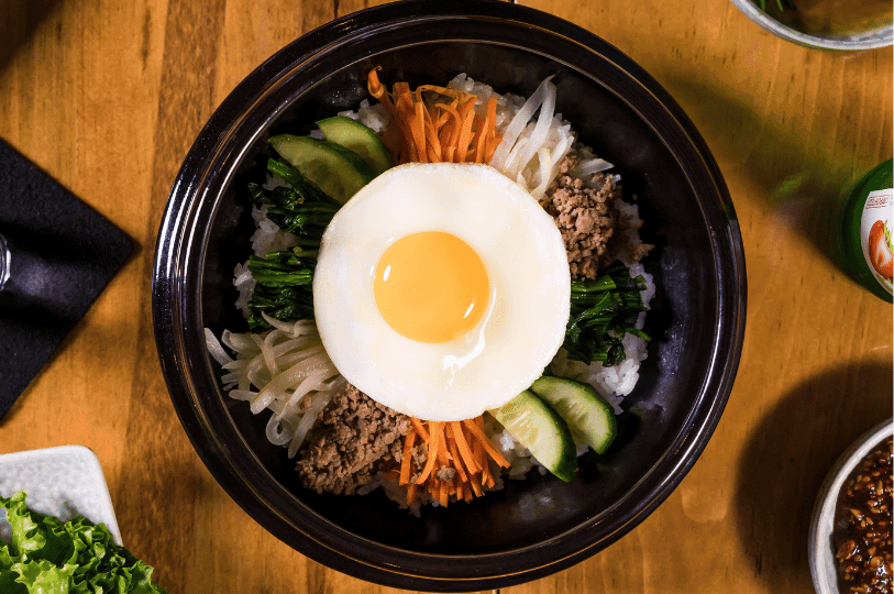 A minimalist, overhead shot of a bibimbap bowl on a wooden table. The ingredients—carrots, spinach, cucumbers, and ground meat—are arranged symmetrically around a central fried egg, creating a clean and aesthetic presentation. A minimalist, overhead shot of a bibimbap bowl on a wooden table. The ingredients—carrots, spinach, cucumbers, and ground meat—are arranged symmetrically around a central fried egg, creating a clean and aesthetic presentation.