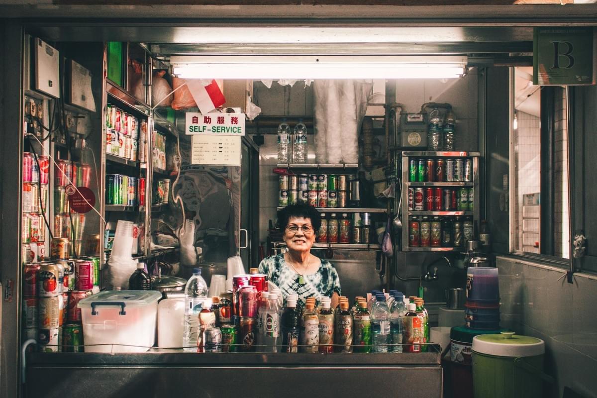 An elderly woman smiles from behind the counter of a small, brightly lit drink stall packed with various bottled and canned beverages. The compact space is neatly organized with shelves of soda and water, featuring a "Self-Service" sign and a small flag overhead. An elderly woman smiles from behind the counter of a small, brightly lit drink stall packed with various bottled and canned beverages. The compact space is neatly organized with shelves of soda and water, featuring a "Self-Service" sign and a small flag overhead.