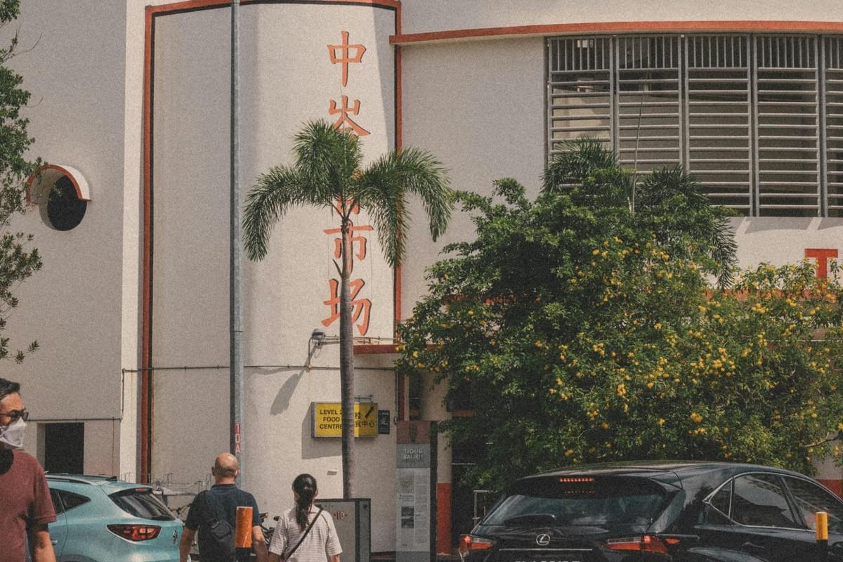 A curved white building facade featuring vertical red Chinese characters rises behind lush green foliage and a tall palm tree. Below, pedestrians walk along the sidewalk near passing cars, while a yellow sign indicates the entrance to a food center on the second level. A curved white building facade featuring vertical red Chinese characters rises behind lush green foliage and a tall palm tree. Below, pedestrians walk along the sidewalk near passing cars, while a yellow sign indicates the entrance to a food center on the second level.