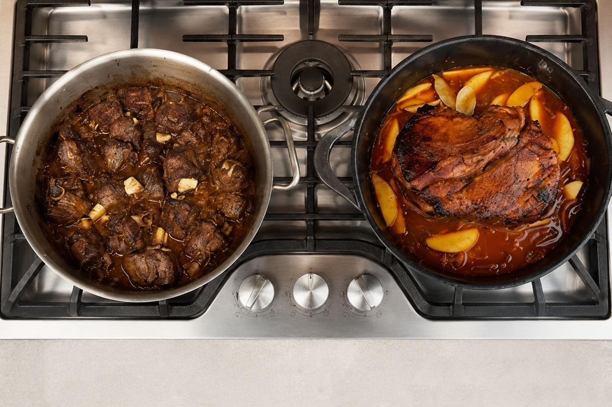 Overhead shot of a gas stovetop with two pots, one containing cubed beef in dark gravy and the other holding a large roast with apples in sauce Overhead shot of a gas stovetop with two pots, one containing cubed beef in dark gravy and the other holding a large roast with apples in sauce