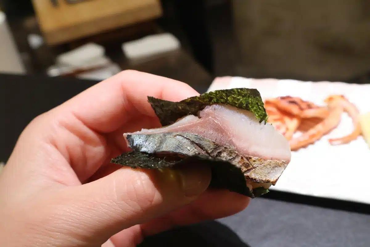 A hand holds a piece of seared fish wrapped in a crisp sheet of dark nori seaweed. In the background, additional dishes are served on a white plate over a dark table. A hand holds a piece of seared fish wrapped in a crisp sheet of dark nori seaweed. In the background, additional dishes are served on a white plate over a dark table.