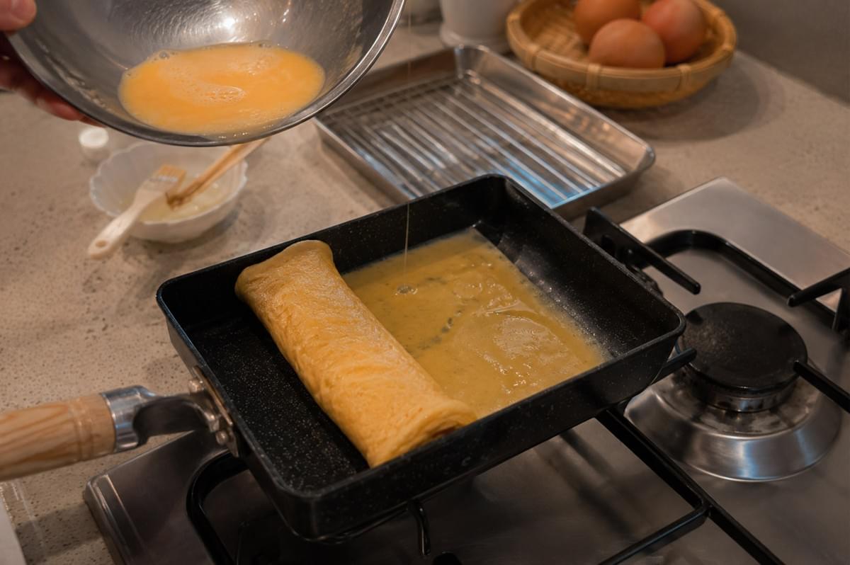 Raw whisked egg mixture is poured from a metal bowl into a rectangular tamagoyaki pan heated on a gas stove burner, spreading beneath an already rolled, golden omelet layer resting at the front edge of the pan. Raw whisked egg mixture is poured from a metal bowl into a rectangular tamagoyaki pan heated on a gas stove burner, spreading beneath an already rolled, golden omelet layer resting at the front edge of the pan.