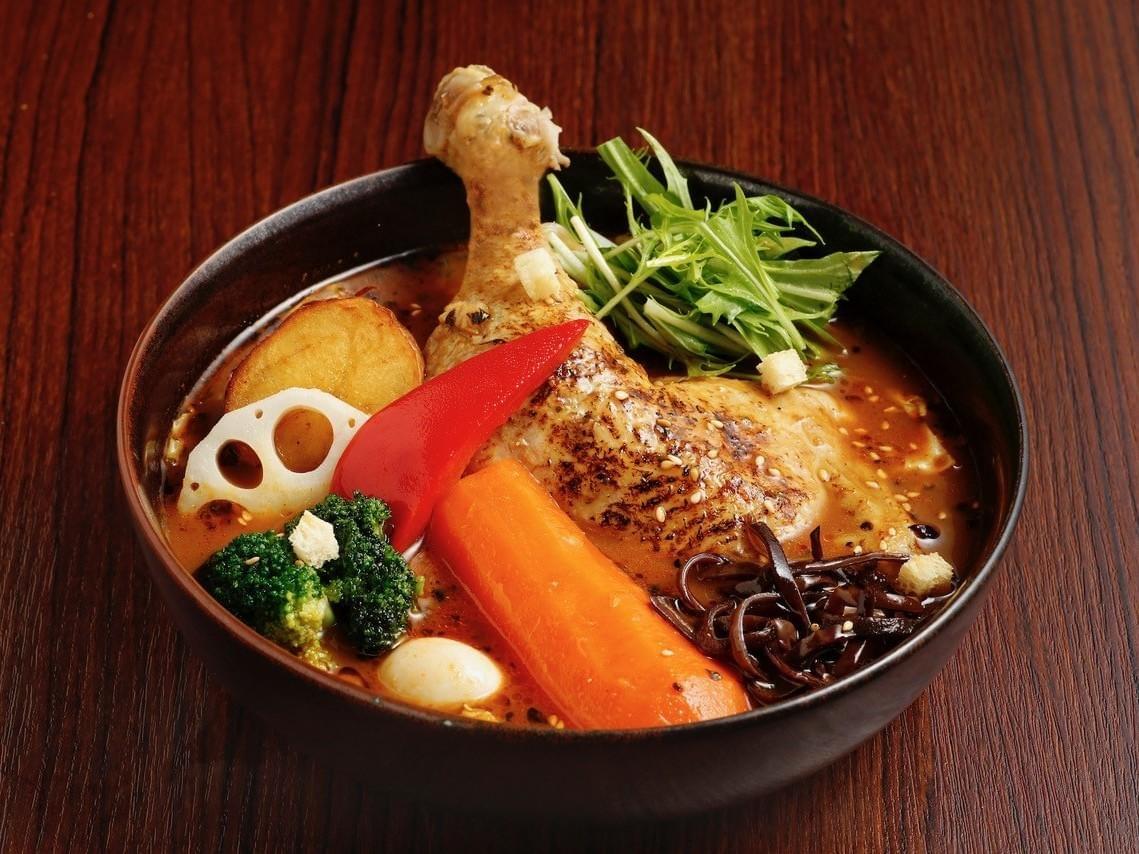 A large bowl of Japanese soup curry features a whole roasted chicken leg nestled alongside a vibrant assortment of vegetables, including a thick carrot chunk, broccoli, red bell pepper, lotus root, and fresh greens. The dark, flavorful broth is sprinkled with sesame seeds and sits in a black ceramic bowl against a rustic wooden background. A large bowl of Japanese soup curry features a whole roasted chicken leg nestled alongside a vibrant assortment of vegetables, including a thick carrot chunk, broccoli, red bell pepper, lotus root, and fresh greens. The dark, flavorful broth is sprinkled with sesame seeds and sits in a black ceramic bowl against a rustic wooden background.