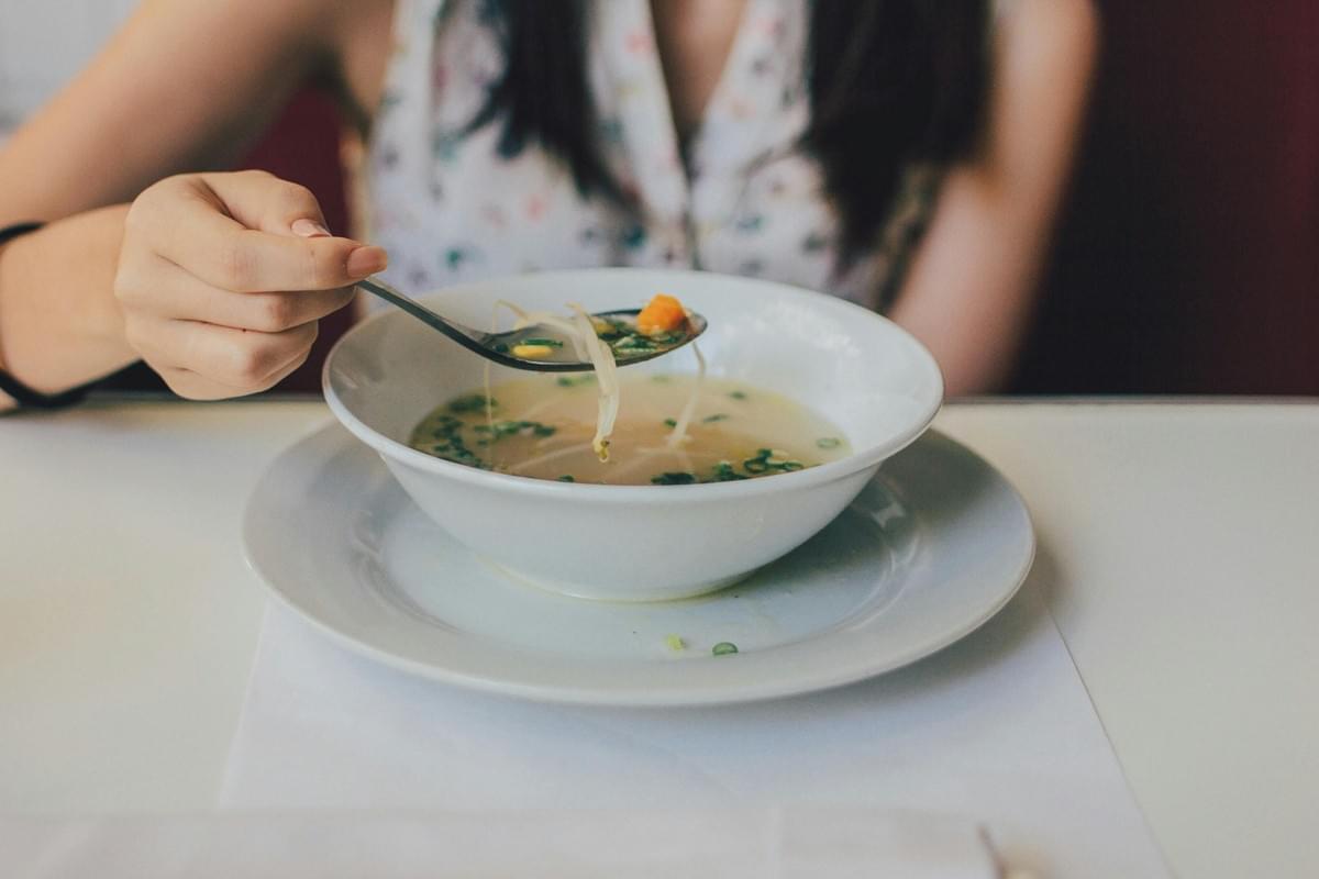 A person holds a spoon filled with soup, noodles, and vegetables over a large white bowl on a matching saucer. The bowl is filled with a clear broth containing green onions and sprouts, set against a clean white tabletop. A person holds a spoon filled with soup, noodles, and vegetables over a large white bowl on a matching saucer. The bowl is filled with a clear broth containing green onions and sprouts, set against a clean white tabletop.