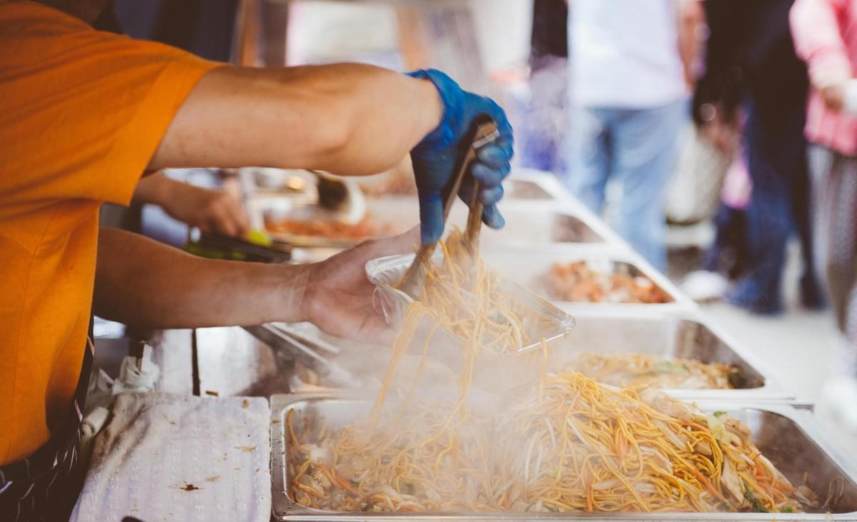A person wearing an orange shirt and a blue glove uses metal tongs to scoop steaming stir-fried noodles into a foil container. The hot noodles are served from large metal catering trays at a busy outdoor food stall or market. A person wearing an orange shirt and a blue glove uses metal tongs to scoop steaming stir-fried noodles into a foil container. The hot noodles are served from large metal catering trays at a busy outdoor food stall or market.