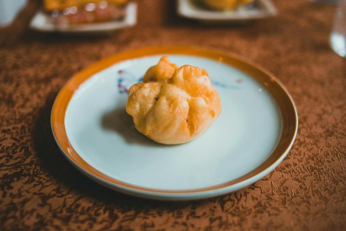A single fried dumpling rests in the center of a white dish with an orange rim. The small plate sits on a patterned brown tablecloth in a warm, dimly lit restaurant setting. A single fried dumpling rests in the center of a white dish with an orange rim. The small plate sits on a patterned brown tablecloth in a warm, dimly lit restaurant setting.