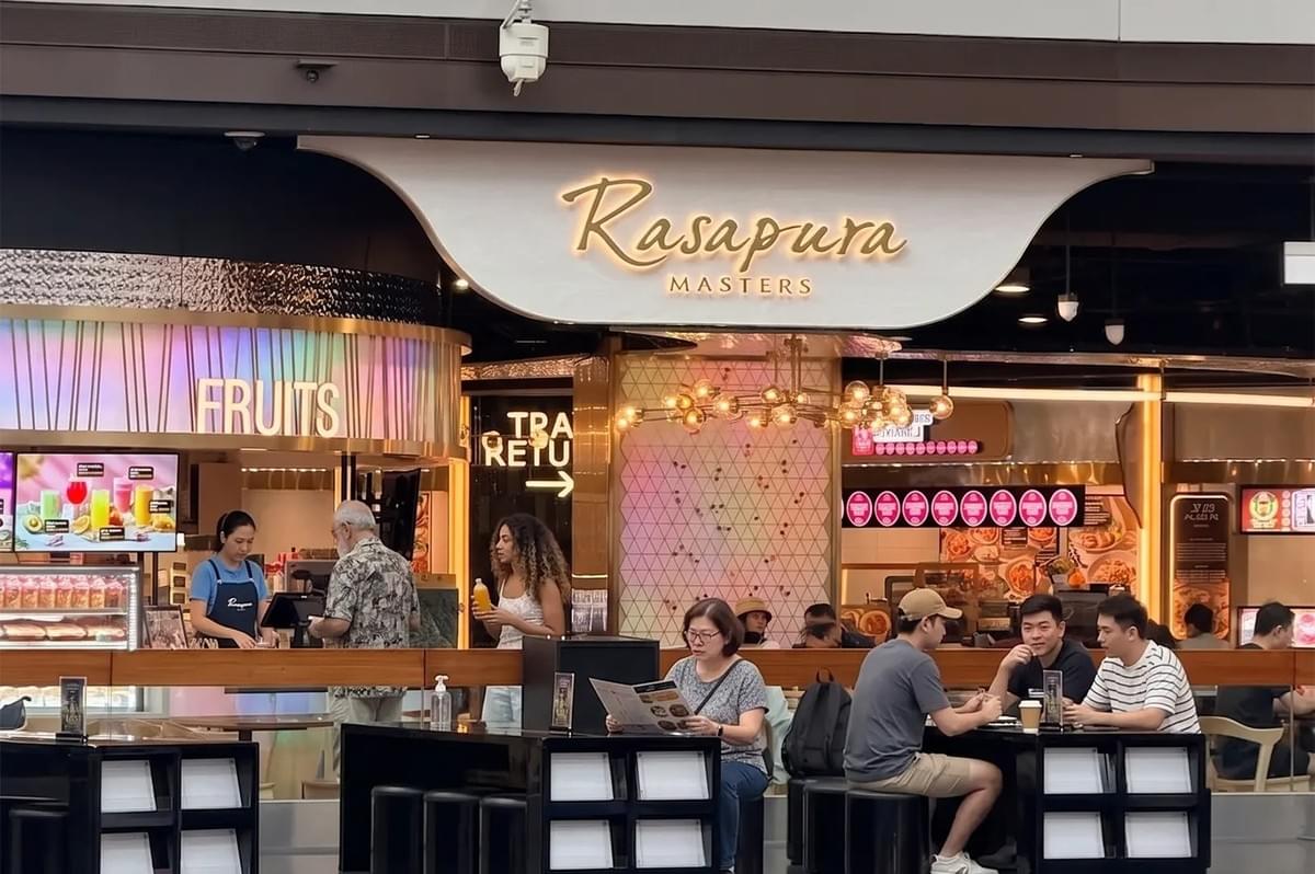Rasapura Masters food court entrance in Singapore with customers dining at tables and a fruit juice stall. Rasapura Masters food court entrance in Singapore with customers dining at tables and a fruit juice stall.