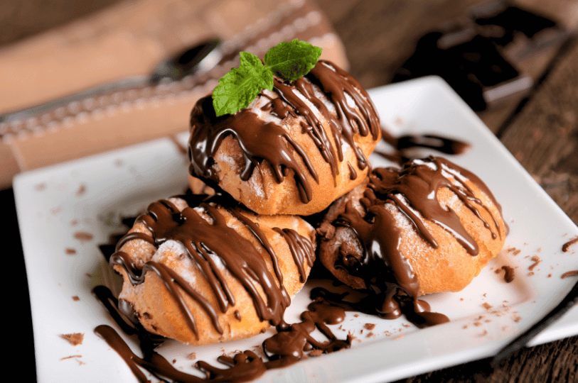 Three profiteroles stacked on a square white plate, dusted lightly with powdered sugar, drizzled with a zig-zag of chocolate sauce, and garnished with a fresh mint leaf. Three profiteroles stacked on a square white plate, dusted lightly with powdered sugar, drizzled with a zig-zag of chocolate sauce, and garnished with a fresh mint leaf.