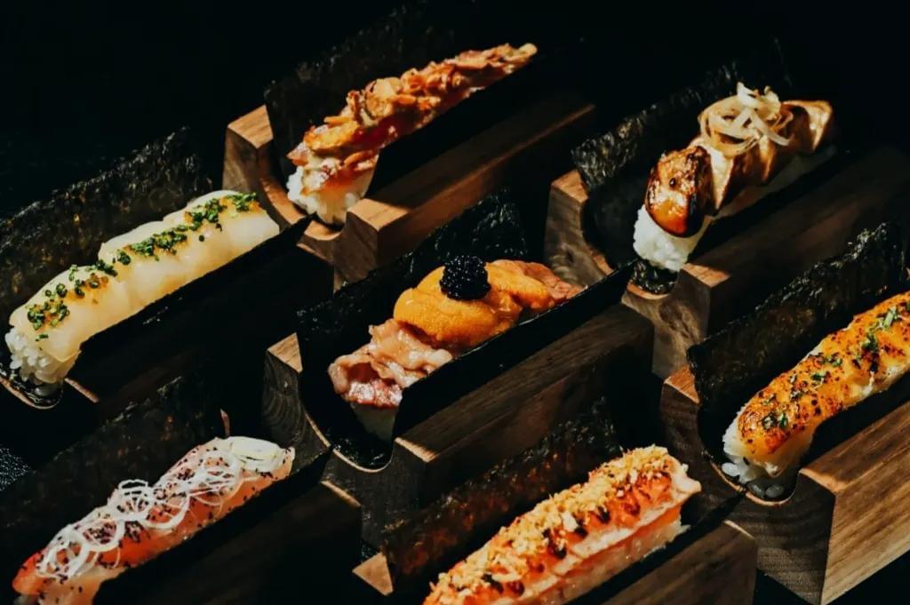 An array of premium open-faced sushi handrolls, featuring toppings like uni with caviar and seared scallops, is presented in individual dark wooden holders. The rolls are elegantly arranged against a black background, highlighting the vibrant colors and textures of the fresh seafood and garnished rice. An array of premium open-faced sushi handrolls, featuring toppings like uni with caviar and seared scallops, is presented in individual dark wooden holders. The rolls are elegantly arranged against a black background, highlighting the vibrant colors and textures of the fresh seafood and garnished rice.