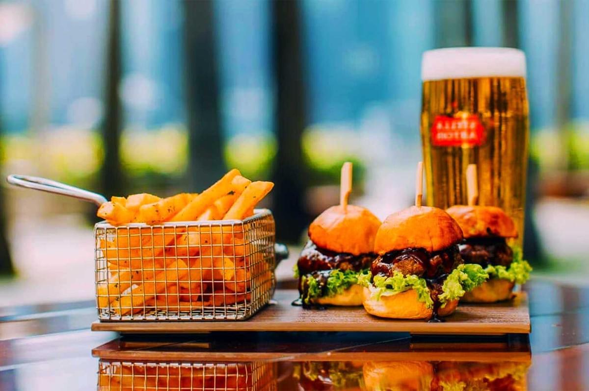 A platter with three beef sliders topped with lettuce and cheese, a basket of crispy fries, and a pint of beer on a wooden table, outdoors. A platter with three beef sliders topped with lettuce and cheese, a basket of crispy fries, and a pint of beer on a wooden table, outdoors.