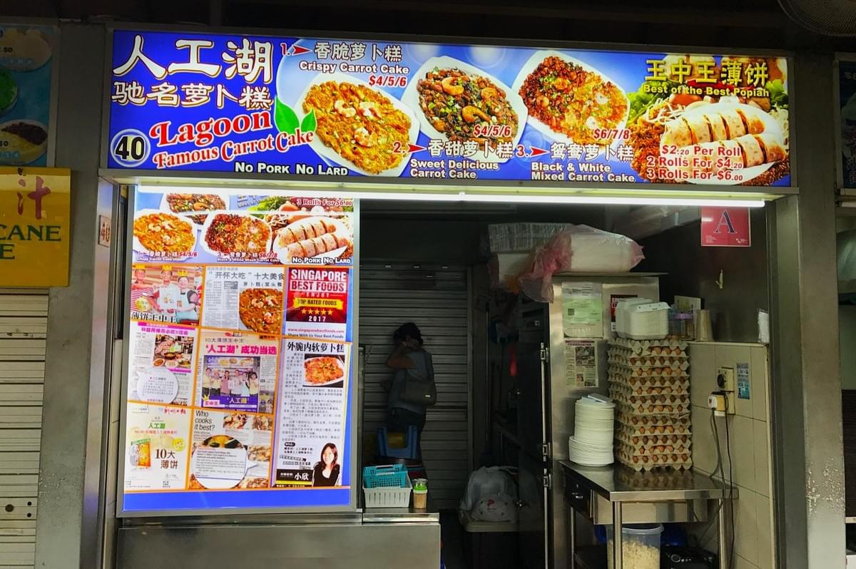 Outdoor food stall with brightly colored signs displaying "Lagoon Famous Carrot Cake." Menu includes crispy, sweet, and mixed carrot cakes. A woman stands inside. Outdoor food stall with brightly colored signs displaying "Lagoon Famous Carrot Cake." Menu includes crispy, sweet, and mixed carrot cakes. A woman stands inside.