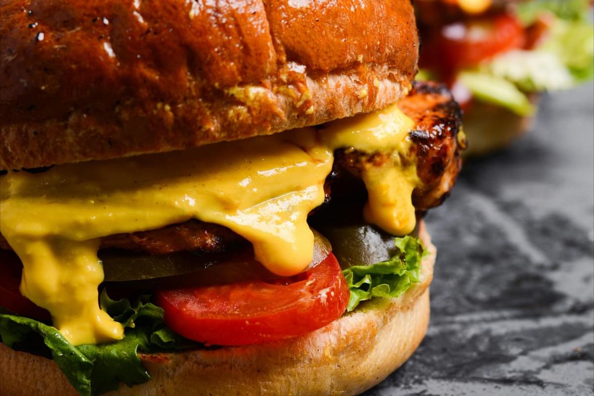 A close-up shot shows melted yellow cheese dripping over a grilled patty, sliced tomatoes, pickles, and fresh lettuce between toasted buns. The background is softly blurred, highlighting the texture of the food resting on a dark, textured surface. A close-up shot shows melted yellow cheese dripping over a grilled patty, sliced tomatoes, pickles, and fresh lettuce between toasted buns. The background is softly blurred, highlighting the texture of the food resting on a dark, textured surface.