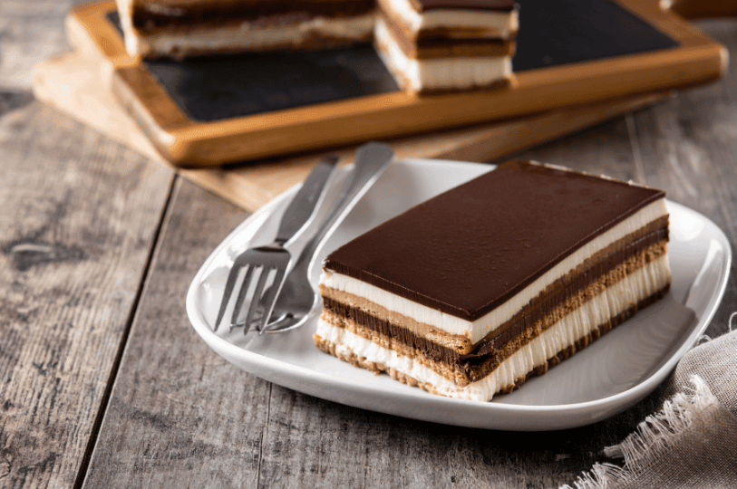 A beautifully layered slice of opera cake highlighting the rich coffee and chocolate tiers, served on a square white plate with two forks against a textured wooden background. A beautifully layered slice of opera cake highlighting the rich coffee and chocolate tiers, served on a square white plate with two forks against a textured wooden background.