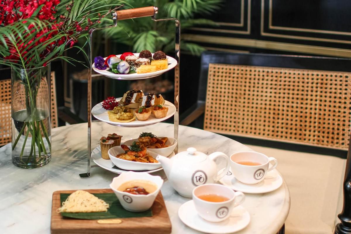 A three-tiered metal tray filled with various sweet and savory high tea delicacies sits on a marble table alongside a white teapot and teacups. Next to the stand, a vase of vibrant red flowers and additional small dessert dishes complete the elegant high tea setting. A three-tiered metal tray filled with various sweet and savory high tea delicacies sits on a marble table alongside a white teapot and teacups. Next to the stand, a vase of vibrant red flowers and additional small dessert dishes complete the elegant high tea setting.