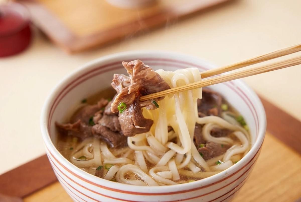 Chopsticks lifting a portion of beef and udon noodles from a steaming bowl of soup. Chopsticks lifting a portion of beef and udon noodles from a steaming bowl of soup.