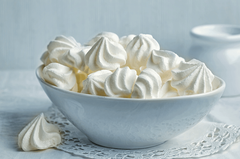 A white ceramic bowl filled to the brim with small, bright white meringue cookies, set against a soft blue background with one stray cookie resting on a lace doily in the foreground. A white ceramic bowl filled to the brim with small, bright white meringue cookies, set against a soft blue background with one stray cookie resting on a lace doily in the foreground.