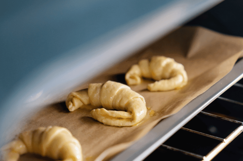 Three raw, hand-rolled croissants sitting on a parchment-lined baking pan inside an oven, emphasizing why delicate, butter-laminated pastries require precise baking temperatures to melt the butter quickly and create perfectly flaky layers. Three raw, hand-rolled croissants sitting on a parchment-lined baking pan inside an oven, emphasizing why delicate, butter-laminated pastries require precise baking temperatures to melt the butter quickly and create perfectly flaky layers.