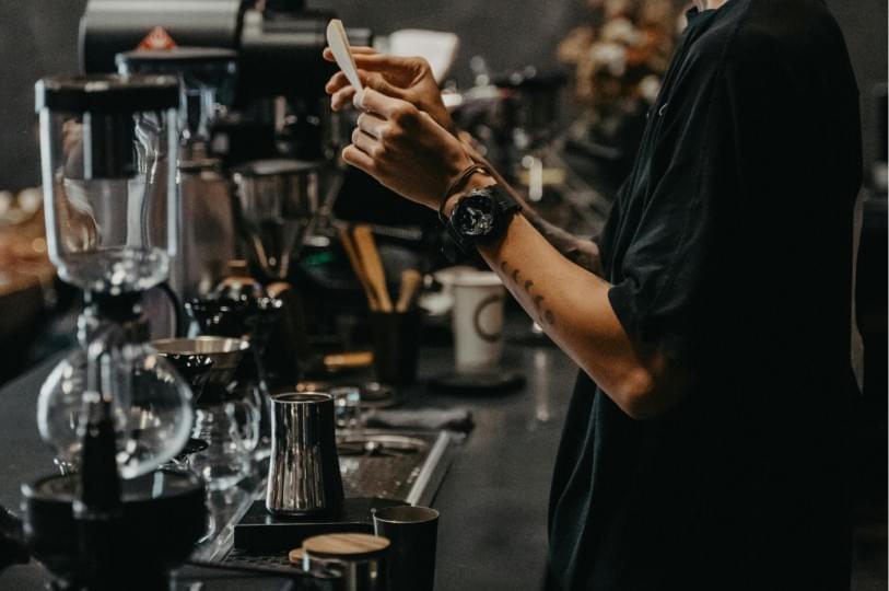 A barista in a black shirt preparing a drink behind a dark, modern coffee bar filled with professional equipment like a siphon brewer and pitchers. A barista in a black shirt preparing a drink behind a dark, modern coffee bar filled with professional equipment like a siphon brewer and pitchers.