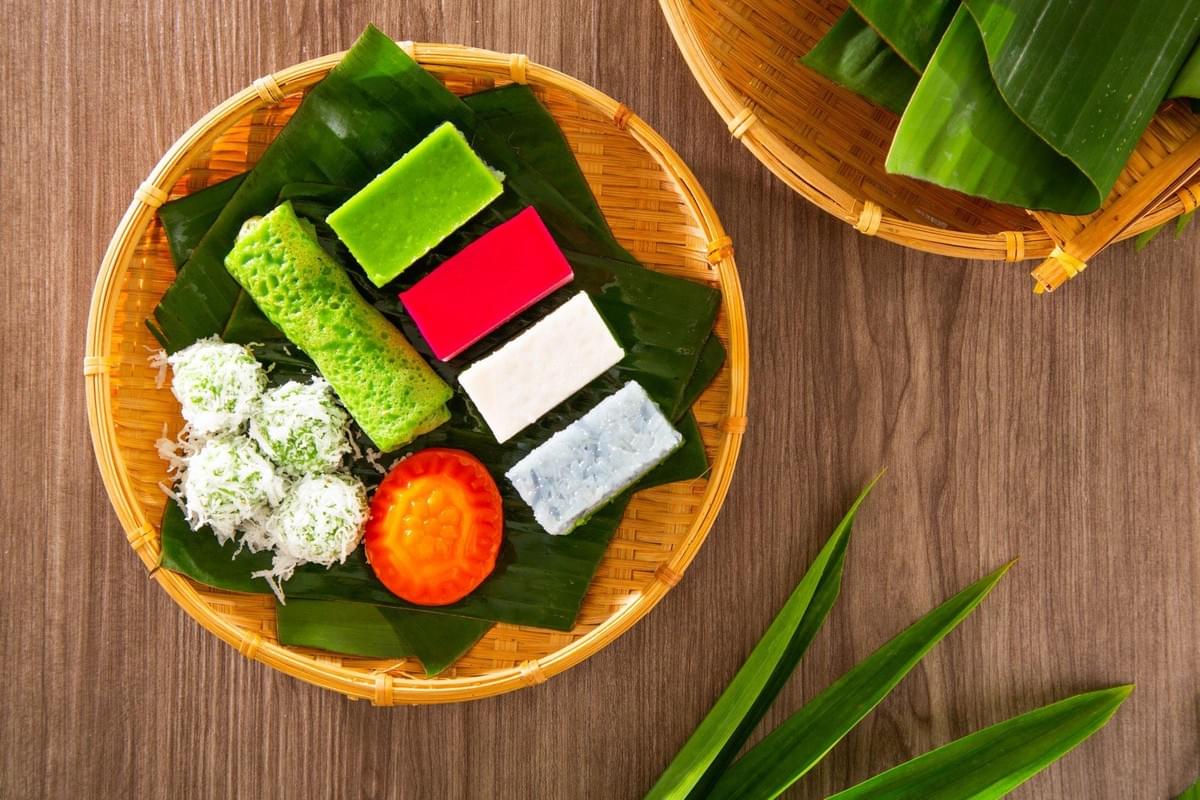 A wicker platter lined with banana leaves features a colorful assortment of traditional Southeast Asian Nyonya kuih sweets, including pandan-flavored rolls, layered cakes, and coconut-coated rice balls. Long green pandan leaves and a basket rest on the wooden tabletop next to the vibrant platter. A wicker platter lined with banana leaves features a colorful assortment of traditional Southeast Asian Nyonya kuih sweets, including pandan-flavored rolls, layered cakes, and coconut-coated rice balls. Long green pandan leaves and a basket rest on the wooden tabletop next to the vibrant platter.