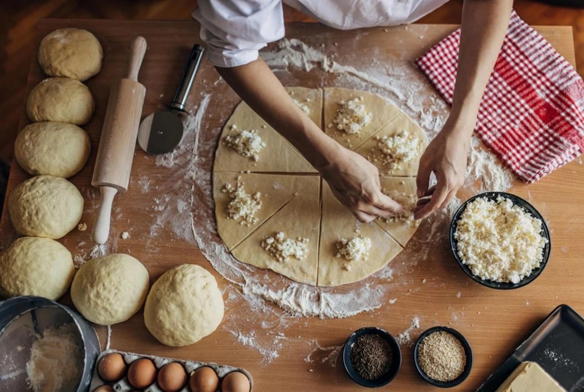 Top-down view of a baker placing cheese filling onto sliced circular dough surrounded by dough balls and baking ingredients. Top-down view of a baker placing cheese filling onto sliced circular dough surrounded by dough balls and baking ingredients.