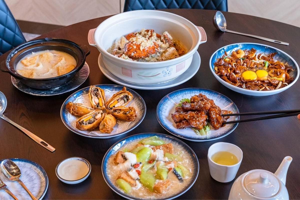 A round wooden dining table is filled with a colorful spread of various Chinese zi char dishes, including a large ceramic casserole of crab, wok-fried hor fun topped with raw egg yolks, glazed ribs, and fried rolls. The table is set with small serving bowls, tea cups, spoons, and chopsticks ready for a shared meal. A round wooden dining table is filled with a colorful spread of various Chinese zi char dishes, including a large ceramic casserole of crab, wok-fried hor fun topped with raw egg yolks, glazed ribs, and fried rolls. The table is set with small serving bowls, tea cups, spoons, and chopsticks ready for a shared meal.
