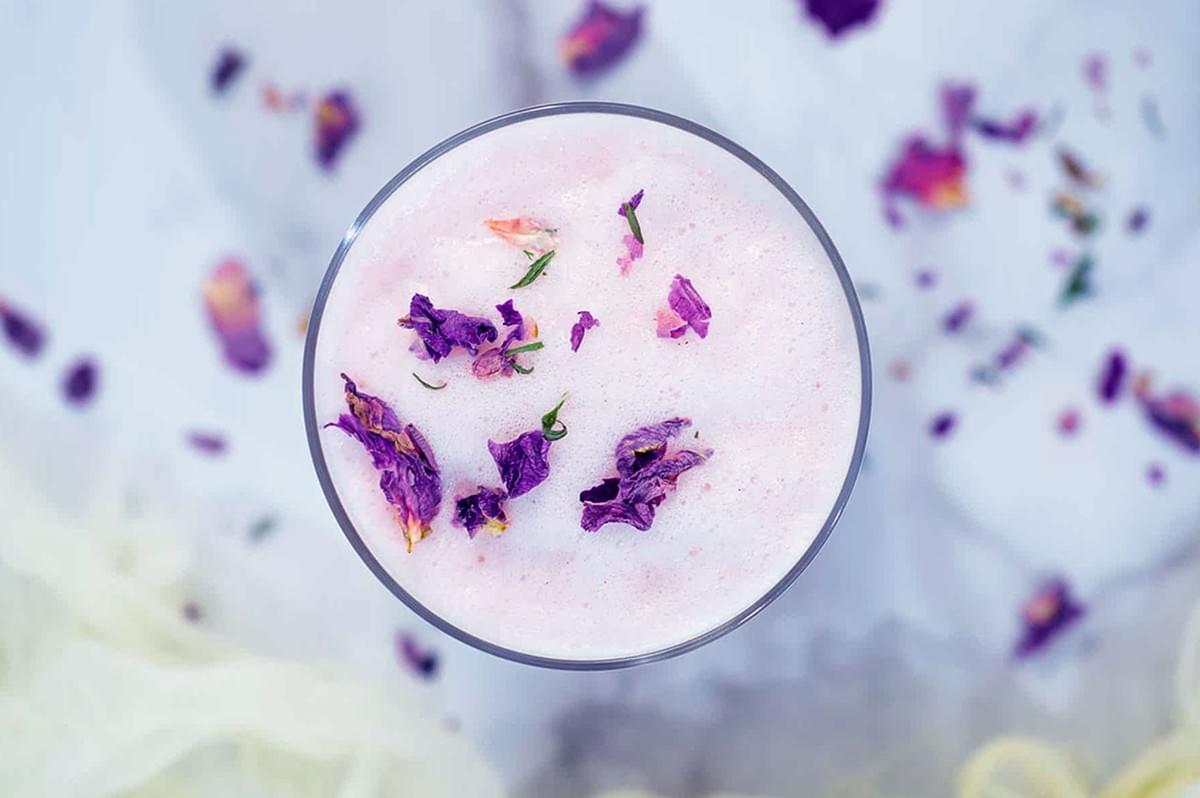 A frothy pink drink in a glass topped with vibrant purple flower petals. The background is soft and blurred with scattered petals, creating a serene, elegant feel. A frothy pink drink in a glass topped with vibrant purple flower petals. The background is soft and blurred with scattered petals, creating a serene, elegant feel.