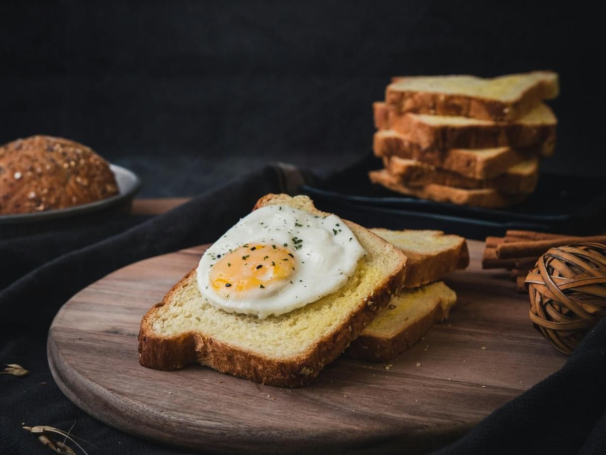 A fried sunny-side-up egg seasoned with herbs sits atop a slice of buttered toast on a wooden cutting board. In the dark, rustic background, a stack of sliced bread and a whole grain roll complete the breakfast setting. A fried sunny-side-up egg seasoned with herbs sits atop a slice of buttered toast on a wooden cutting board. In the dark, rustic background, a stack of sliced bread and a whole grain roll complete the breakfast setting.