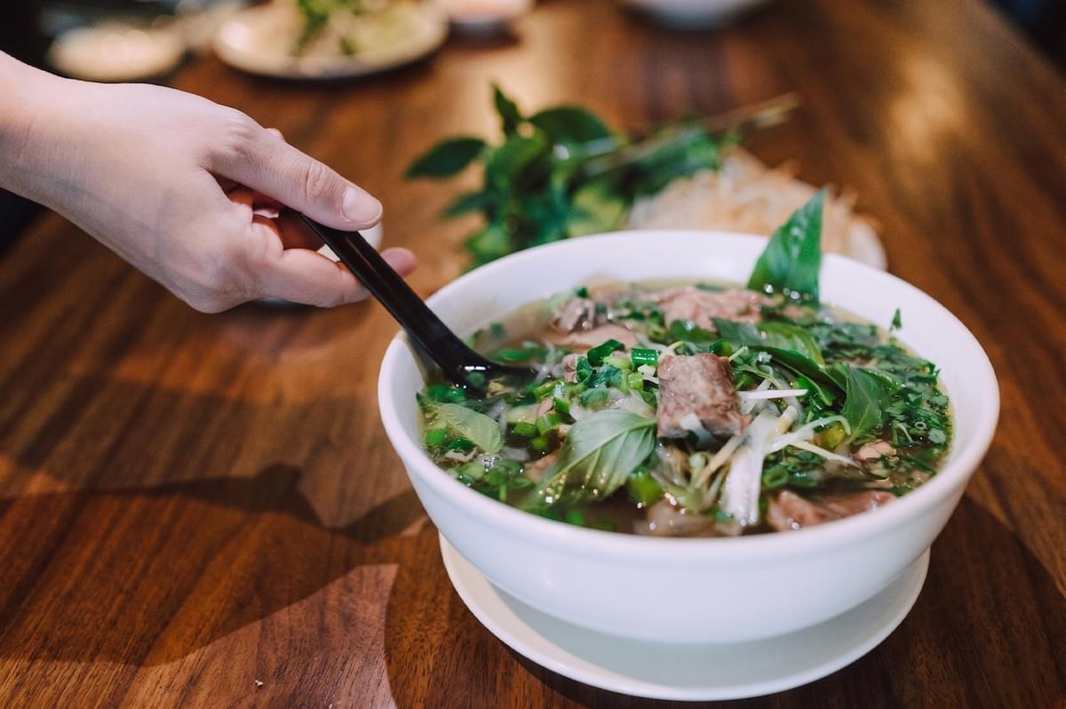 Close-up of a steaming bowl of beef noodle soup filled with herbs and green onions on a wooden table. Close-up of a steaming bowl of beef noodle soup filled with herbs and green onions on a wooden table.