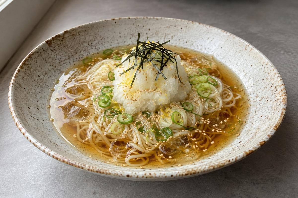 A specked ceramic bowl contains a chilled noodle dish submerged in a light amber broth with ice cubes, piled high with a mound of finely grated white radish, sprinkled with toasted sesame seeds, chopped scallions, yellow yuzu zest, and thin strips of nori seaweed. A specked ceramic bowl contains a chilled noodle dish submerged in a light amber broth with ice cubes, piled high with a mound of finely grated white radish, sprinkled with toasted sesame seeds, chopped scallions, yellow yuzu zest, and thin strips of nori seaweed.