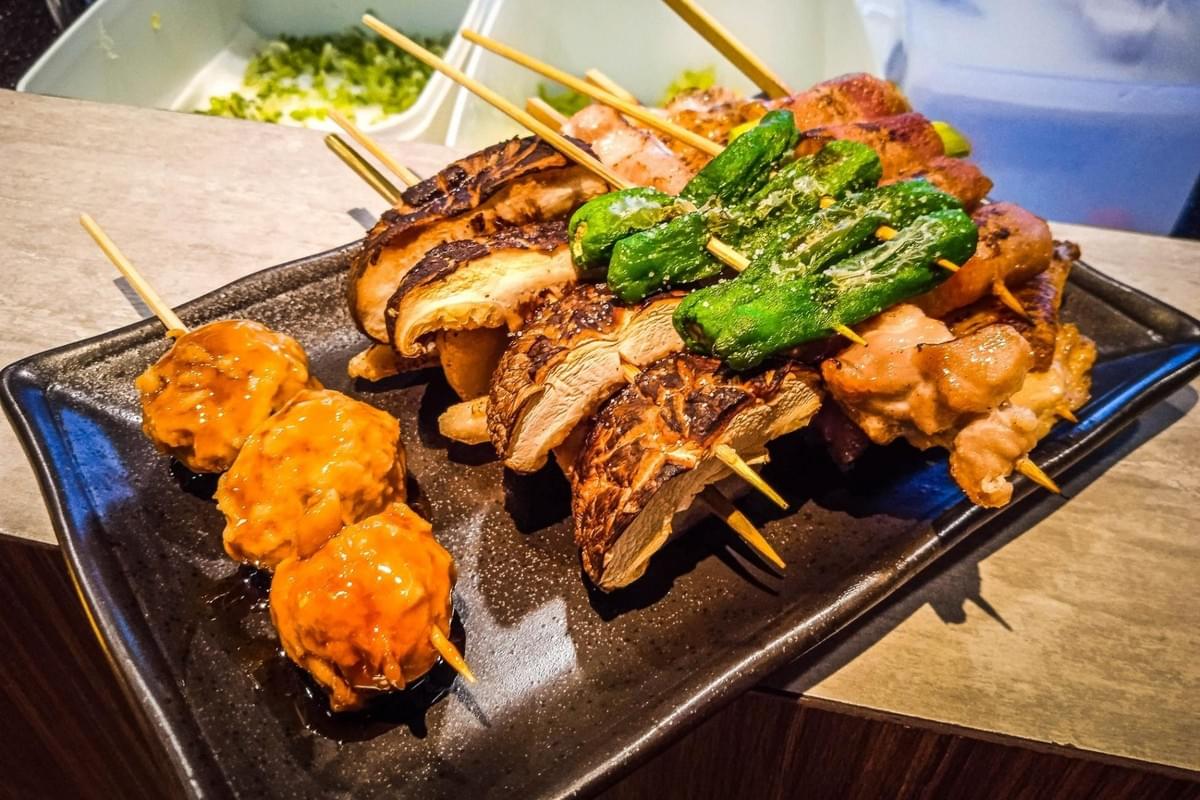 An assortment of freshly grilled Japanese yakitori skewers served on a dark rectangular plate. The platter includes glazed meatball skewers, sliced shiitake mushrooms, and blistered shishito peppers. An assortment of freshly grilled Japanese yakitori skewers served on a dark rectangular plate. The platter includes glazed meatball skewers, sliced shiitake mushrooms, and blistered shishito peppers.