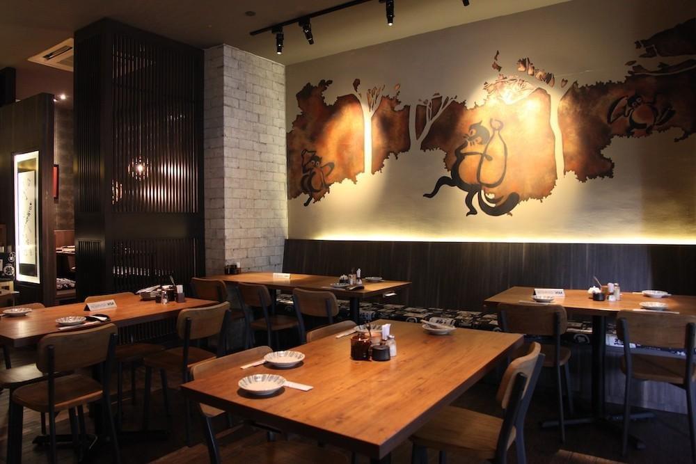 A dimly lit Japanese restaurant dining room features wooden tables set with plates, chopsticks, and condiment jars. Above the banquette seating, a large illuminated mural depicts whimsical tanuki characters along a warm, earth-toned wall. A dimly lit Japanese restaurant dining room features wooden tables set with plates, chopsticks, and condiment jars. Above the banquette seating, a large illuminated mural depicts whimsical tanuki characters along a warm, earth-toned wall.