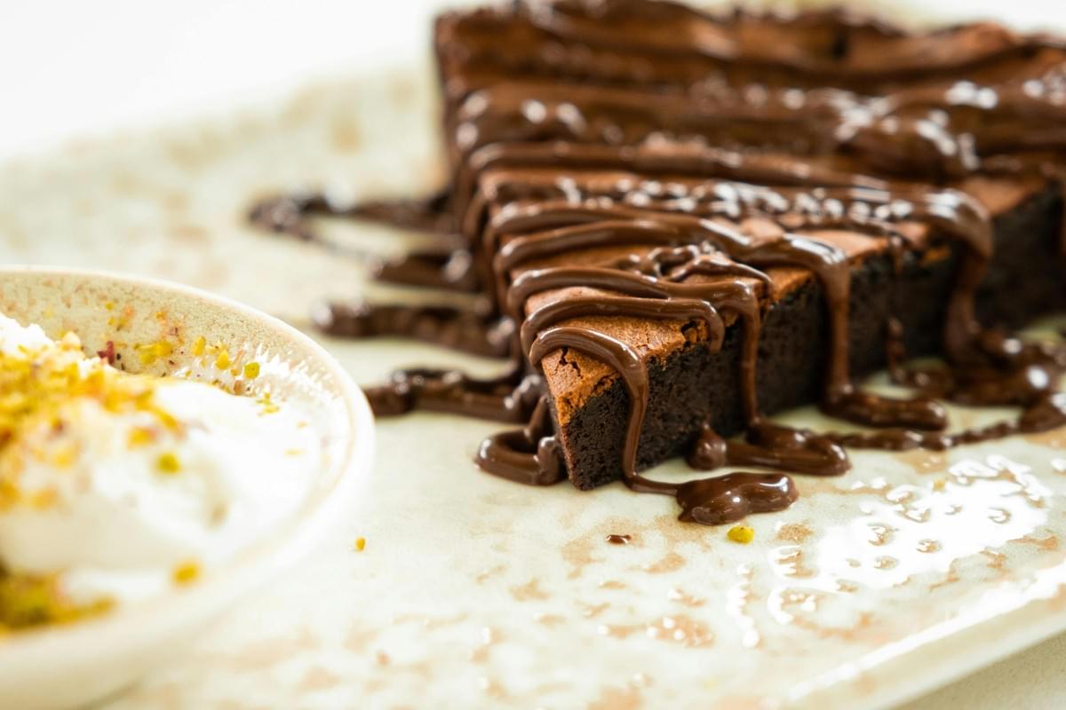A wedge of rich chocolate brownie cake sits on a speckled plate, generously drizzled with thick chocolate sauce. Next to the dessert, a small white bowl holds a scoop of vanilla ice cream topped with crushed green pistachios. A wedge of rich chocolate brownie cake sits on a speckled plate, generously drizzled with thick chocolate sauce. Next to the dessert, a small white bowl holds a scoop of vanilla ice cream topped with crushed green pistachios.
