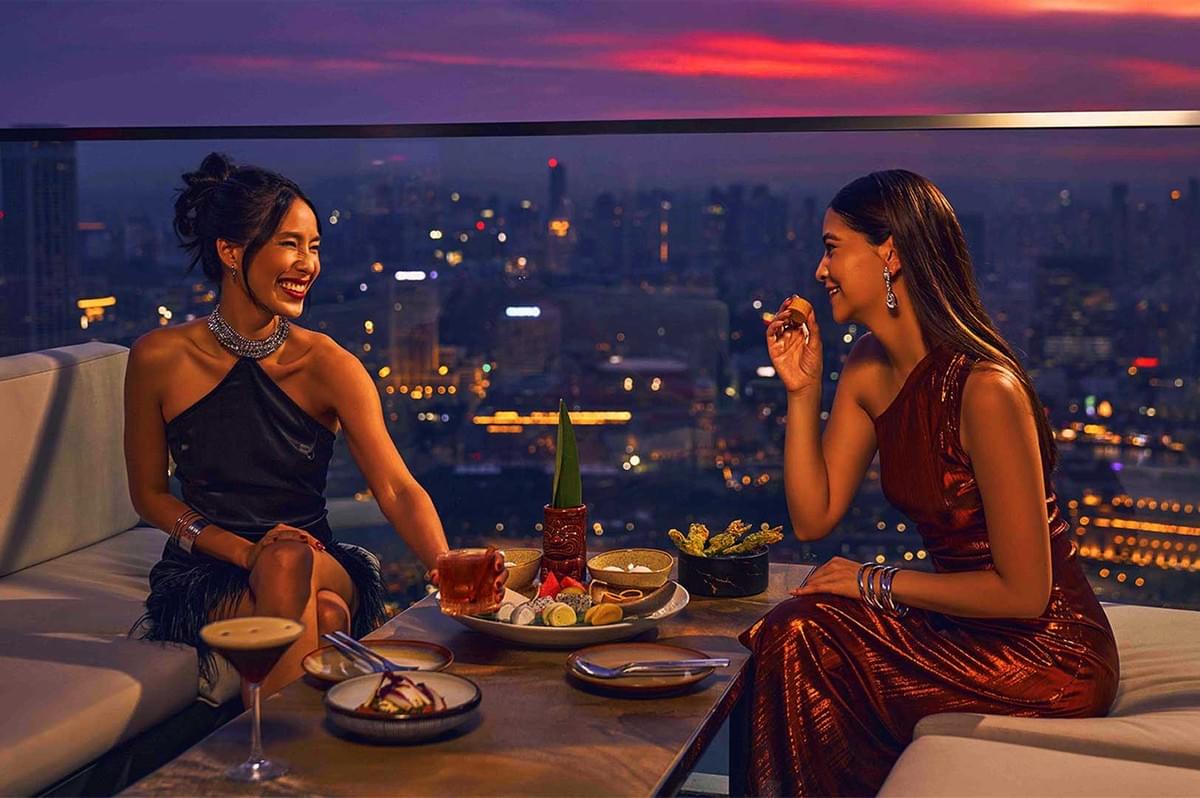 Two women elegantly dressed enjoy cocktails and snacks on a rooftop at sunset, with a vibrant cityscape in the background. They sit, smiling and laughing, creating a joyful and lively atmosphere. Two women elegantly dressed enjoy cocktails and snacks on a rooftop at sunset, with a vibrant cityscape in the background. They sit, smiling and laughing, creating a joyful and lively atmosphere.