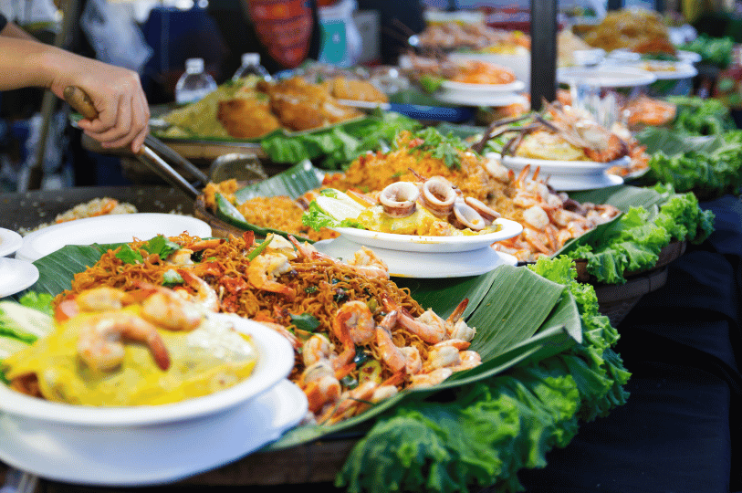 A bustling Thai street food stall display with various dishes on banana leaves, including stir-fried noodles with shrimp, calamari, and assorted seafood delicacies. A bustling Thai street food stall display with various dishes on banana leaves, including stir-fried noodles with shrimp, calamari, and assorted seafood delicacies.