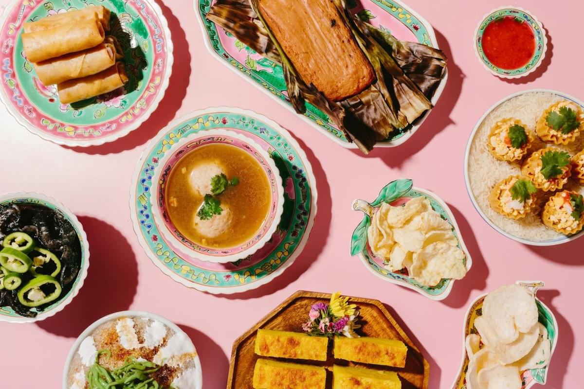 An overhead flat-lay shot features a colorful spread of traditional Peranakan and Southeast Asian dishes arranged on decorative ceramic plates against a bright pink backdrop. The array includes spring rolls, meatball soup, grilled otah wrapped in banana leaves, pie tee shells filled with savory ingredients, and iced dessert. An overhead flat-lay shot features a colorful spread of traditional Peranakan and Southeast Asian dishes arranged on decorative ceramic plates against a bright pink backdrop. The array includes spring rolls, meatball soup, grilled otah wrapped in banana leaves, pie tee shells filled with savory ingredients, and iced dessert.