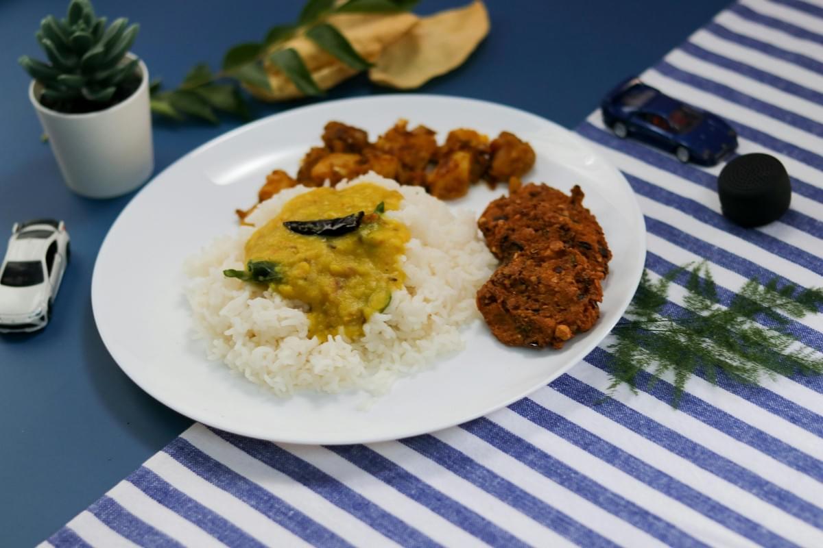 A white plate on a blue and white striped cloth holds a serving of white rice topped with yellow dal, accompanied by a side of spiced potatoes and crispy fritters. The background features a small potted succulent, green leaves, and two toy cars scattered on a solid blue surface. A white plate on a blue and white striped cloth holds a serving of white rice topped with yellow dal, accompanied by a side of spiced potatoes and crispy fritters. The background features a small potted succulent, green leaves, and two toy cars scattered on a solid blue surface.