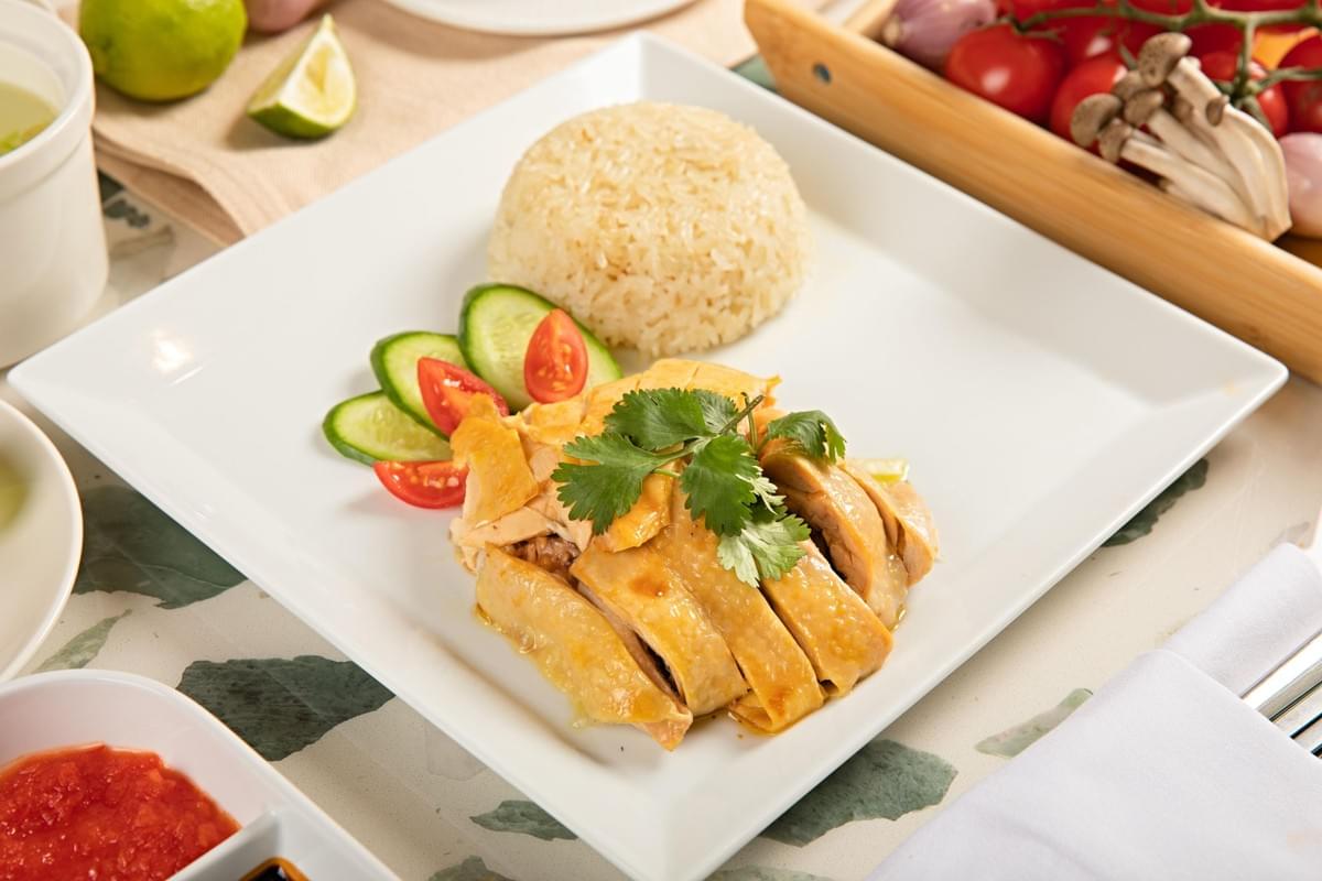 In the image, a serving of Hainanese chicken rice is neatly arranged on a square white plate, featuring a dome of seasoned rice alongside sliced poached chicken topped with fresh cilantro. The dish is garnished with cucumber slices and cherry tomatoes, surrounded by dipping sauces and raw ingredients in the background. In the image, a serving of Hainanese chicken rice is neatly arranged on a square white plate, featuring a dome of seasoned rice alongside sliced poached chicken topped with fresh cilantro. The dish is garnished with cucumber slices and cherry tomatoes, surrounded by dipping sauces and raw ingredients in the background.