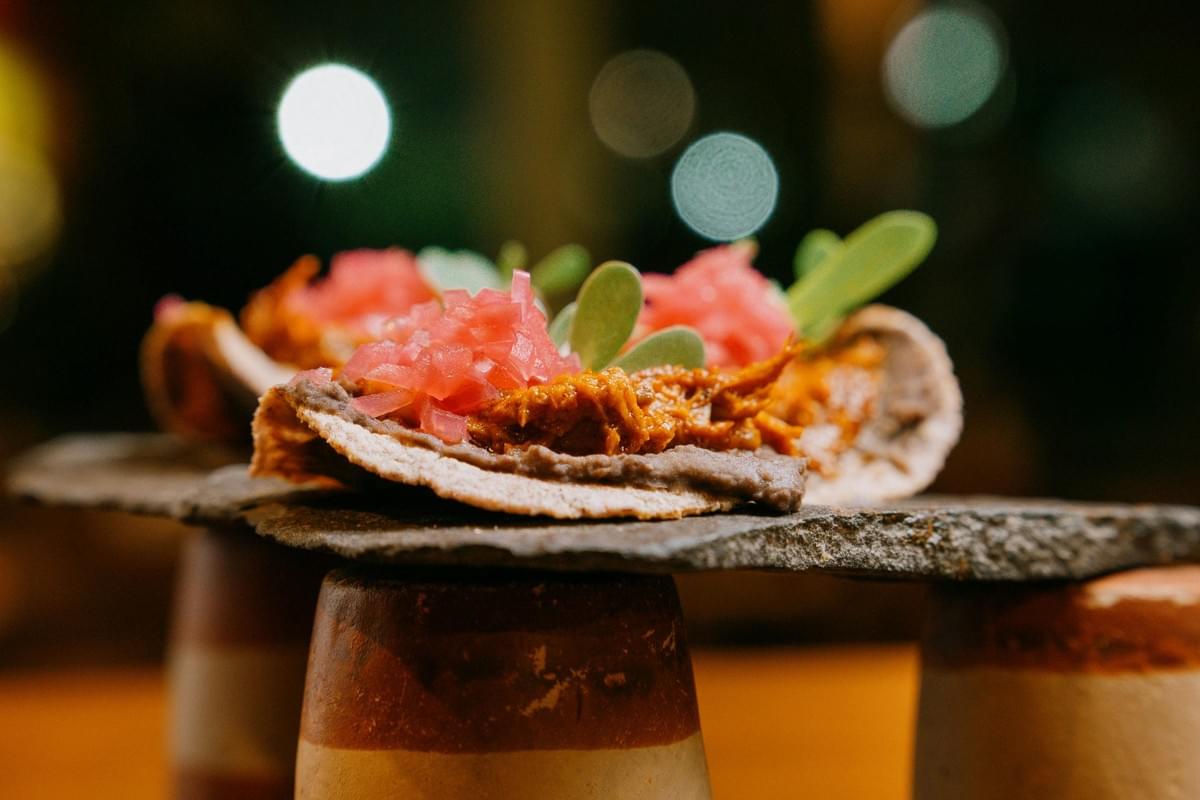 A close-up shot showcases Yucatecan panuchos or tlacoyos topped with black bean paste, shredded meat, pickled red onions, and fresh microgreens, elegantly served on a flat slate stone. The slate is balanced atop inverted clay cups against a warm, softly blurred background with ambient bokeh lights. A close-up shot showcases Yucatecan panuchos or tlacoyos topped with black bean paste, shredded meat, pickled red onions, and fresh microgreens, elegantly served on a flat slate stone. The slate is balanced atop inverted clay cups against a warm, softly blurred background with ambient bokeh lights.